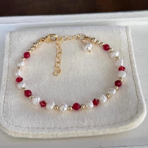 May include: A delicate gold bracelet featuring white pearls, red beads, and gold accents. The bracelet is displayed on a white surface. The clasp is gold and adjustable.