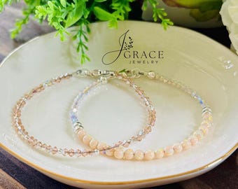 May include: Two delicate bracelets resting on a white dish. One bracelet features small, light-colored, iridescent round beads, while the other consists of faceted, light-brown crystal beads. Both include silver-toned metal clasps and accents.