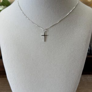 May include: A delicate silver-toned necklace featuring a simple, slender cross pendant suspended from a thin, polished chain. The metal has a bright, reflective finish, and the cross design is minimalist with clean, straight edges and a smooth surface.