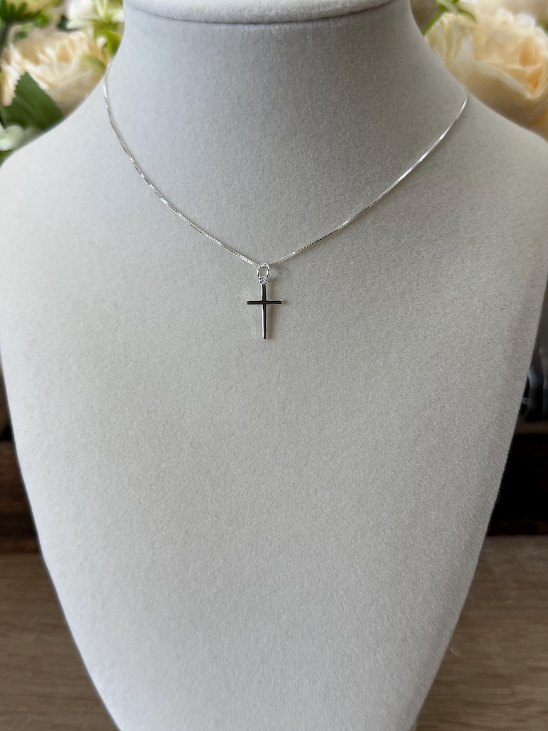 May include: A delicate silver-toned necklace featuring a simple, slender cross pendant suspended from a thin, polished chain. The metal has a bright, reflective finish, and the cross design is minimalist with clean, straight edges and a smooth surface.