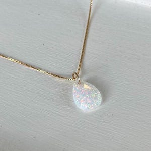 May include: A gold-colored chain necklace with a pear-shaped pendant. The pendant is white with iridescent flecks, giving it a shimmering appearance. The necklace is displayed on a white surface.