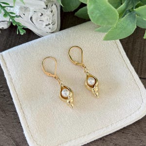 May include: A pair of gold-colored earrings with a pearl accent. The earrings feature a unique design, with a teardrop-shaped setting that cradles a single pearl. The earrings are displayed on a white cloth.