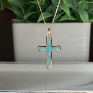 May include: A gold-colored necklace with a cross pendant. The cross has a light blue center and a gold-colored border. The necklace is hanging in front of a blurred background of green plants.