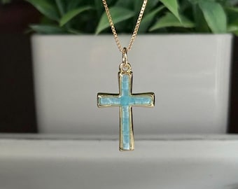 May include: A gold-colored necklace with a cross pendant. The cross has a light blue center and a gold-colored border. The necklace is hanging in front of a blurred background of green plants.