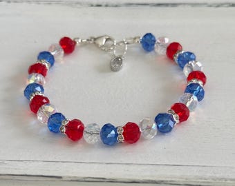 Puede incluir: Un brazalete de cristal rojo, blanco y azul con un cierre plateado. El brazalete está hecho con pequeñas cuentas facetadas.