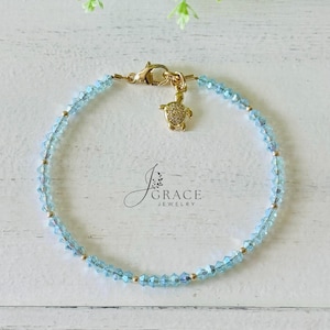 May include: A delicate bracelet featuring light blue faceted crystal beads interspersed with small gold-toned metal spheres. The piece includes a gold-toned lobster clasp and a small turtle-shaped charm with a textured surface. The text J Grace Jewelry is visible.