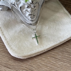 May include: A silver-toned cross pendant suspended from a delicate box chain necklace. The cross features a smooth, polished finish and is attached to the chain by a small circular jump ring. The necklace is presented against a soft, light-colored fabric.