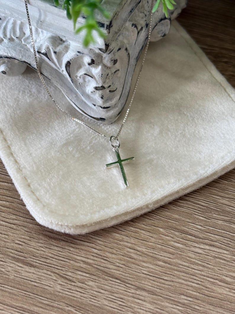 May include: A silver-toned cross pendant suspended from a delicate box chain necklace. The cross features a smooth, polished finish and is attached to the chain by a small circular jump ring. The necklace is presented against a soft, light-colored fabric.