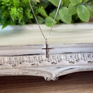 May include: A delicate silver-toned necklace featuring a simple, polished cross pendant suspended from a thin, fine-link chain. The metal has a bright, reflective finish, and the cross design is clean, slender, and minimalist in its overall appearance.