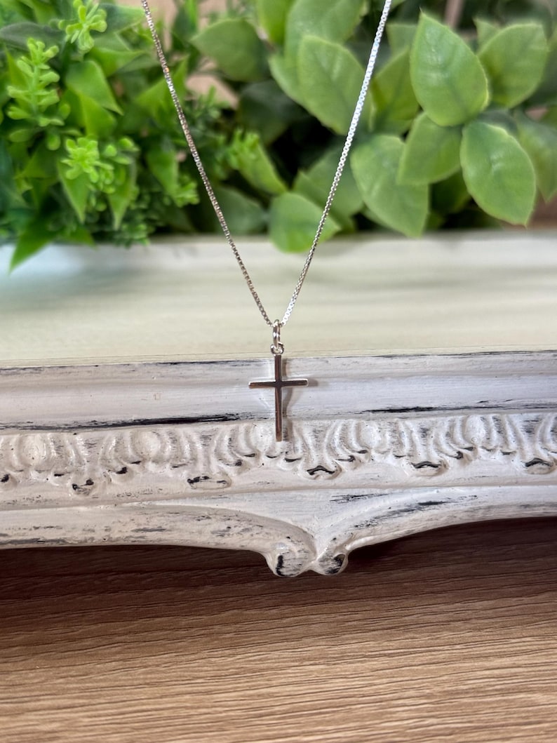 May include: A delicate silver-toned necklace featuring a simple, polished cross pendant suspended from a thin, fine-link chain. The metal has a bright, reflective finish, and the cross design is clean, slender, and minimalist in its overall appearance.