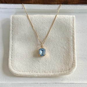 May include: A gold-colored necklace with a rectangular, light blue gemstone pendant. The pendant is set in a gold frame and hangs from a delicate gold chain. The necklace rests on a white, fabric-covered surface.