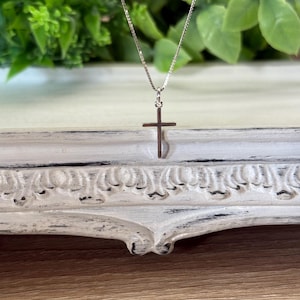 May include: A delicate silver-toned necklace featuring a thin box chain and a small, plain cross pendant. The metal has a polished, reflective finish, and the cross is suspended from a simple jump ring attached to the center of the chain.