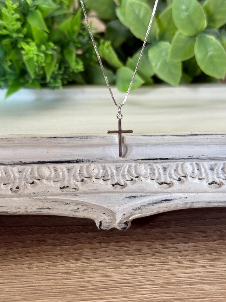 May include: A delicate silver-toned necklace featuring a thin box chain and a small, plain cross pendant. The metal has a polished, reflective finish, and the cross is suspended from a simple jump ring attached to the center of the chain.