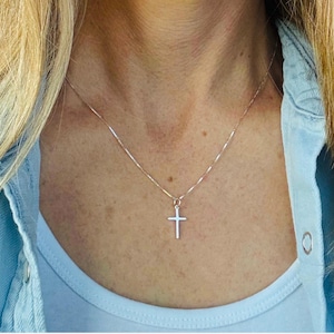 May include: A delicate silver-toned metal chain necklace featuring a small, simple cross pendant. The thin, polished chain rests against the neck, supporting the minimalist cross charm which hangs centered at the base of the neckline.