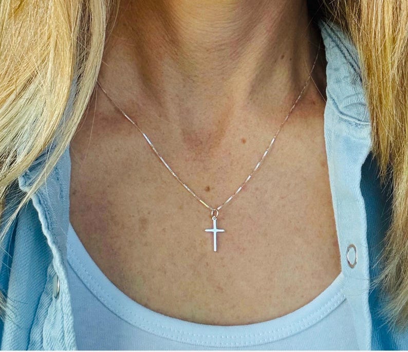May include: A delicate silver-toned metal chain necklace featuring a small, simple cross pendant. The thin, polished chain rests against the neck, supporting the minimalist cross charm which hangs centered at the base of the neckline.
