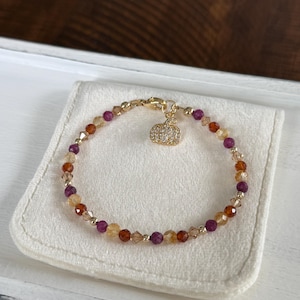 May include: A gold-toned bracelet with a pumpkin charm. The bracelet features a mix of orange, purple, and clear beads. The pumpkin charm is encrusted with small, clear crystals. The bracelet is displayed on a white, rectangular surface.