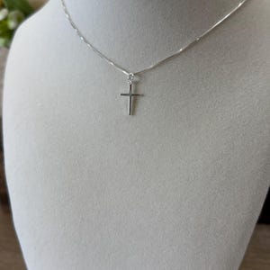 May include: A delicate silver-toned necklace featuring a small, plain cross pendant suspended from a thin, polished box chain. The metal has a bright, reflective finish, and the cross is centered on the chain, creating a simple and clean design.