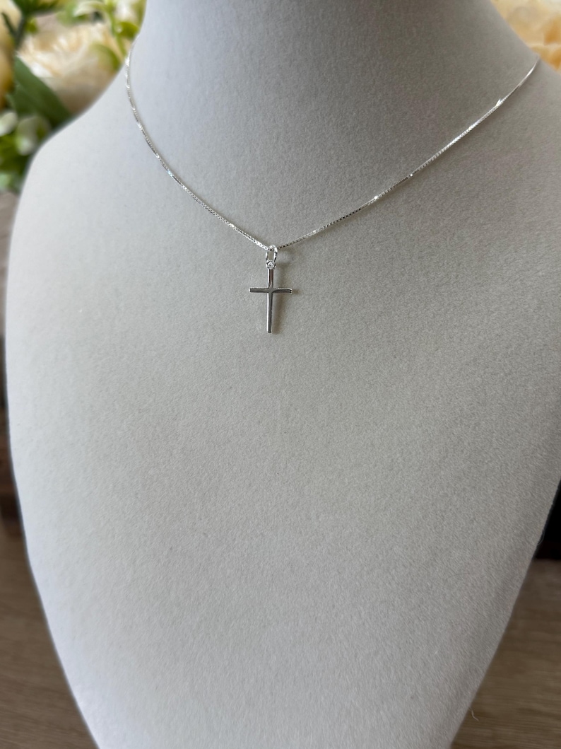 May include: A delicate silver-toned necklace featuring a small, plain cross pendant suspended from a thin, polished box chain. The metal has a bright, reflective finish, and the cross is centered on the chain, creating a simple and clean design.