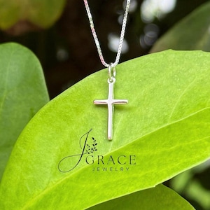 May include: A silver cross pendant suspended from a delicate box chain. The pendant features a smooth, polished finish. The text J GRACE JEWELRY is printed in black on the surface below the cross. The design is simple, featuring a vertical bar.