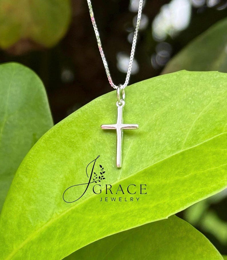 May include: A silver cross pendant suspended from a delicate box chain. The pendant features a smooth, polished finish. The text J GRACE JEWELRY is printed in black on the surface below the cross. The design is simple, featuring a vertical bar.