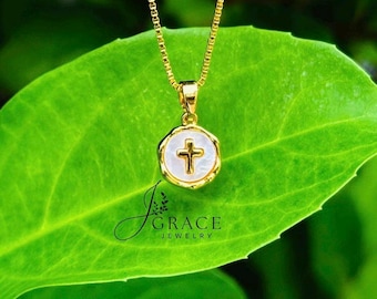 May include: A gold chain necklace featuring a circular pendant with a white mother-of-pearl inlay and a centered gold cross. The pendant is suspended from a delicate box chain. The text J GRACE JEWELRY is visible in black lettering below the pendant.