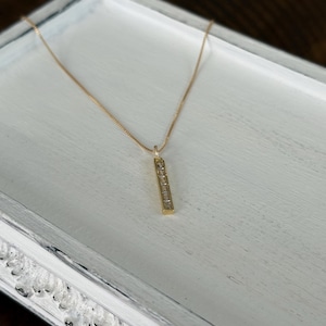 May include: A gold-colored necklace with a rectangular pendant. The pendant is adorned with several small, clear stones. The necklace is displayed on a white, distressed wooden surface.