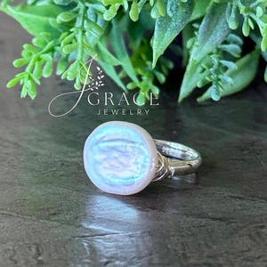 May include: A silver ring featuring a round, iridescent white coin pearl set on a polished band. The band is marked with 925, indicating sterling silver. The text J GRACE JEWELRY is visible above the ring, which is set against a dark surface.