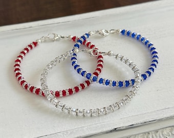 Puede incluir: Tres pulseras de cuentas rojas, transparentes y azules. Cada pulsera presenta pequeñas cuentas espaciadas uniformemente ensartadas en una cadena plateada. Las pulseras se exhiben en una bandeja blanca ornamentada, con un fondo de madera borroso.