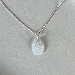 May include: A silver necklace with a pear-shaped, iridescent white pendant. The pendant is faceted, catching the light and displaying a range of colors. The chain is a delicate, silver box chain. The necklace is displayed on a white surface.
