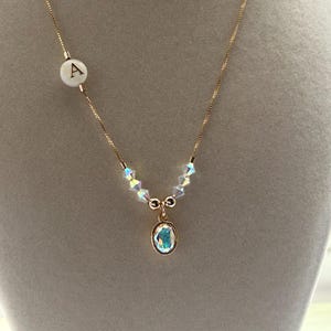 May include: A gold chain necklace with a white bead with the letter 'A' on it. The necklace has a small, oval-shaped, iridescent blue crystal pendant.