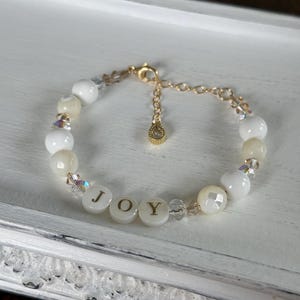 May include: White beaded bracelet with gold accents and the word "JOY" spelled out in gold letters on white beads.