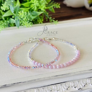 May include: Two delicate bracelets featuring light pink and iridescent crystal beads with silver-toned metal accents and lobster claw clasps. The jewelry is branded with the text J Grace Jewelry. The beads have a faceted finish and a soft, reflective sheen.