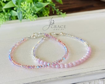 May include: Two delicate bracelets featuring light pink and iridescent crystal beads with silver-toned metal accents and lobster claw clasps. The jewelry is branded with the text J Grace Jewelry. The beads have a faceted finish and a soft, reflective sheen.