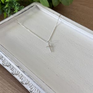 May include: A delicate silver-toned chain necklace featuring a small, simple cross pendant. The thin metal chain connects to the top of the cross, which has a smooth, polished finish and a classic, straight-edged design with no additional embellishments.