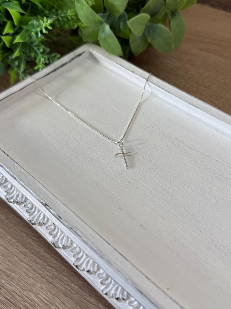 May include: A delicate silver-toned chain necklace featuring a small, simple cross pendant. The thin metal chain connects to the top of the cross, which has a smooth, polished finish and a classic, straight-edged design with no additional embellishments.