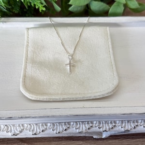 May include: A delicate silver-toned necklace featuring a simple, slender cross pendant suspended from a thin, polished chain. The cross has clean, straight lines and a smooth, reflective metallic finish, resting against a soft, off-white fabric pouch.