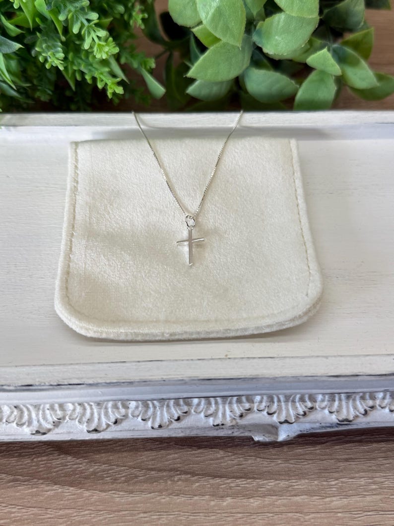 May include: A delicate silver-toned necklace featuring a simple, slender cross pendant suspended from a thin, polished chain. The cross has clean, straight lines and a smooth, reflective metallic finish, resting against a soft, off-white fabric pouch.
