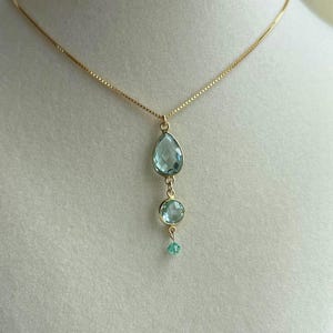 May include: A gold-toned necklace featuring a pendant with three light blue gemstones. The top gemstone is a teardrop shape, followed by a round stone, and a small bead at the bottom. The chain is delicate and hangs on a white background.