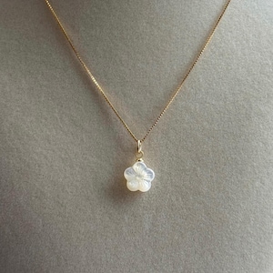 May include: A delicate gold necklace featuring a five-petal flower pendant. The pendant is white and has a subtle, pearlescent sheen. The chain is a fine, gold-colored link, and the necklace is displayed against a neutral background.