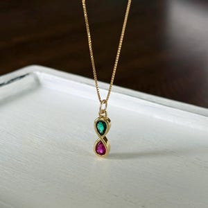May include: A gold-colored necklace with a pendant featuring two teardrop-shaped gemstones, one green and one pink. The gemstones are set in a gold-colored frame, and the necklace is displayed on a white surface.