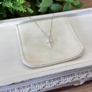 May include: A delicate silver-toned necklace featuring a small, simple cross pendant suspended from a thin, reflective chain. The metal has a polished finish that catches the light, creating a clean and understated appearance for this jewelry piece.