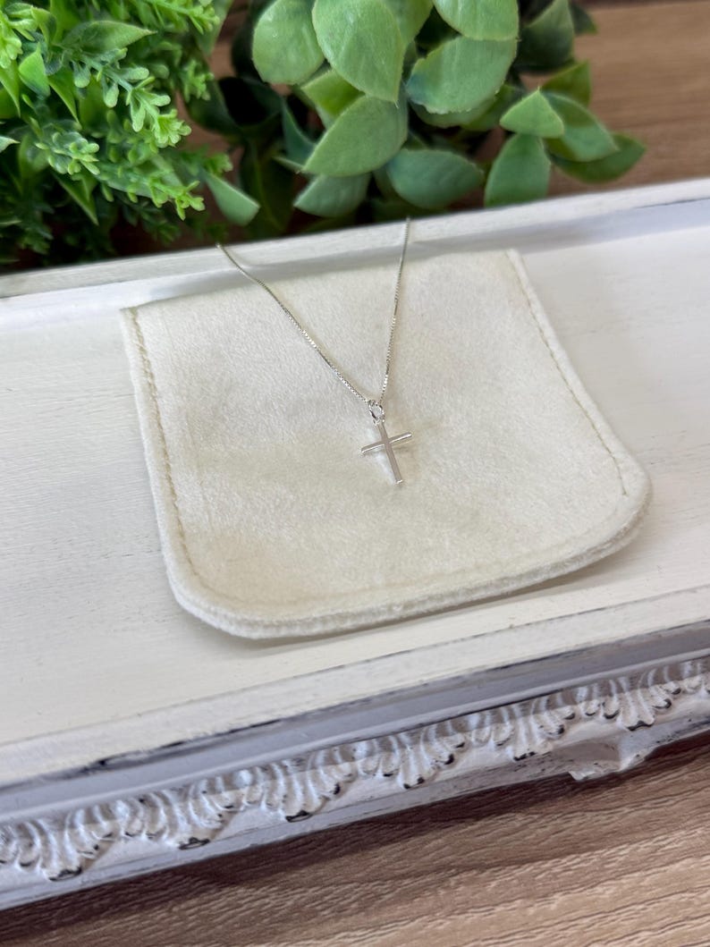 May include: A delicate silver-toned necklace featuring a small, simple cross pendant suspended from a thin, reflective chain. The metal has a polished finish that catches the light, creating a clean and understated appearance for this jewelry piece.