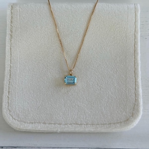 May include: A gold necklace with a rectangular, light blue gemstone pendant. The pendant is set in a gold frame and hangs from a delicate gold chain. The necklace is displayed on a white, folded fabric.
