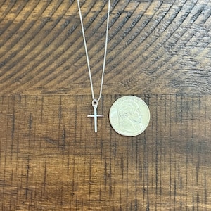 May include: A thin silver chain necklace with a small, plain silver cross pendant. The pendant is positioned next to a United States quarter coin for scale, which has a diameter of approximately 0.955 inches, to show the size of the cross.