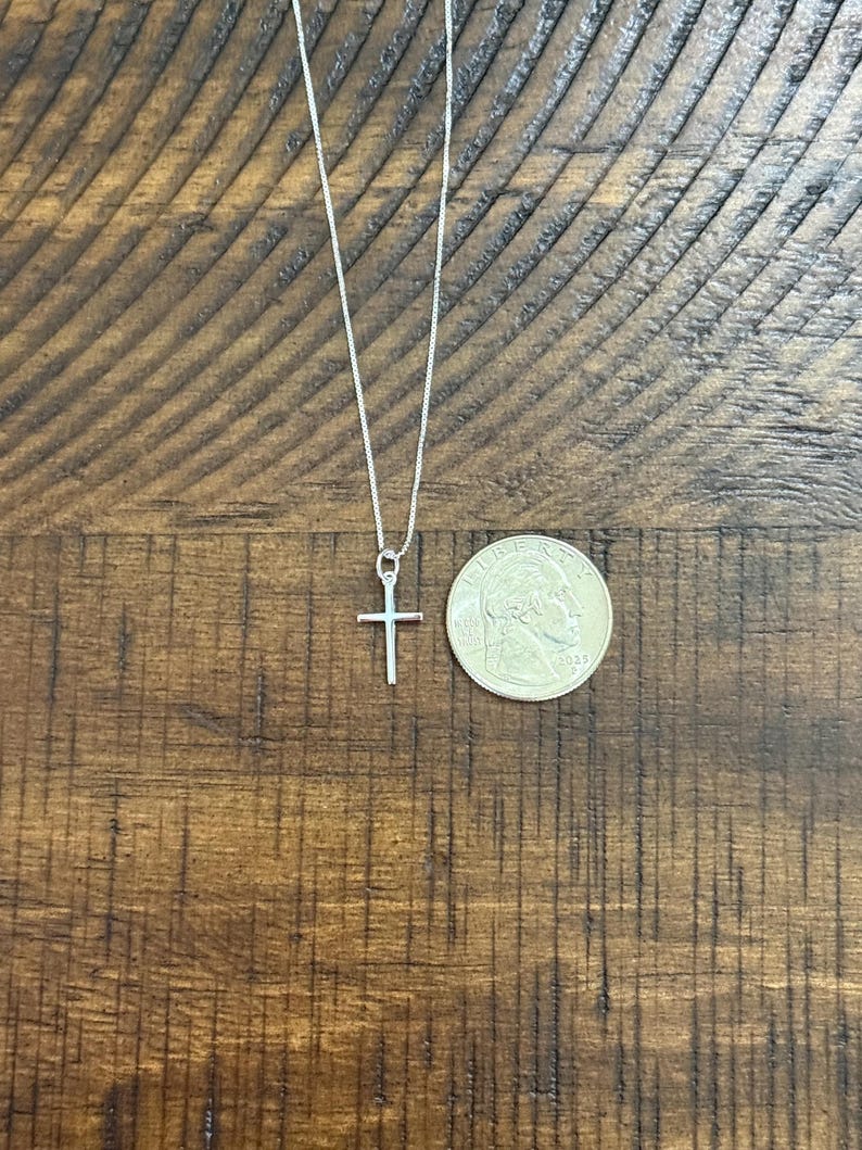 May include: A thin silver chain necklace with a small, plain silver cross pendant. The pendant is positioned next to a United States quarter coin for scale, which has a diameter of approximately 0.955 inches, to show the size of the cross.