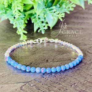May include: A delicate bracelet featuring a strand of small, round, light blue opal-like beads with iridescent flashes, accented by clear crystal beads and a silver-toned metal lobster claw clasp. The jewelry piece is branded with the text J GRACE JEWELRY.