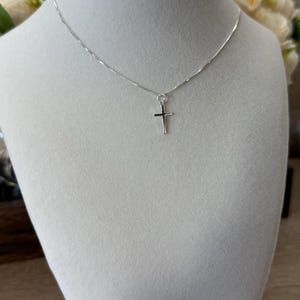 May include: A silver-toned necklace featuring a delicate box chain and a small, simple cross pendant. The metal has a polished, reflective finish. The cross is suspended from a small jump ring attached directly to the thin, metallic chain.