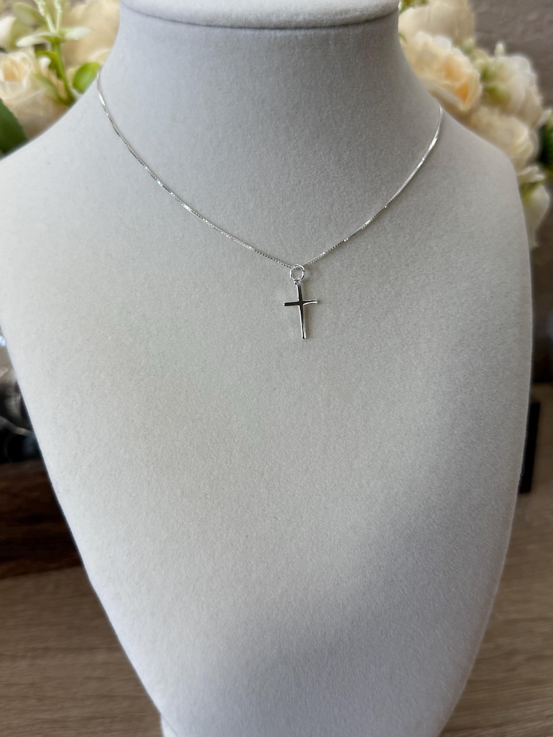 May include: A silver-toned necklace featuring a delicate box chain and a small, simple cross pendant. The metal has a polished, reflective finish. The cross is suspended from a small jump ring attached directly to the thin, metallic chain.