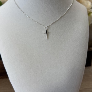 May include: A silver-toned necklace featuring a thin, delicate chain and a small, simple cross pendant. The metal has a polished, reflective finish, and the cross is suspended from a circular jump ring attached to the center of the chain.