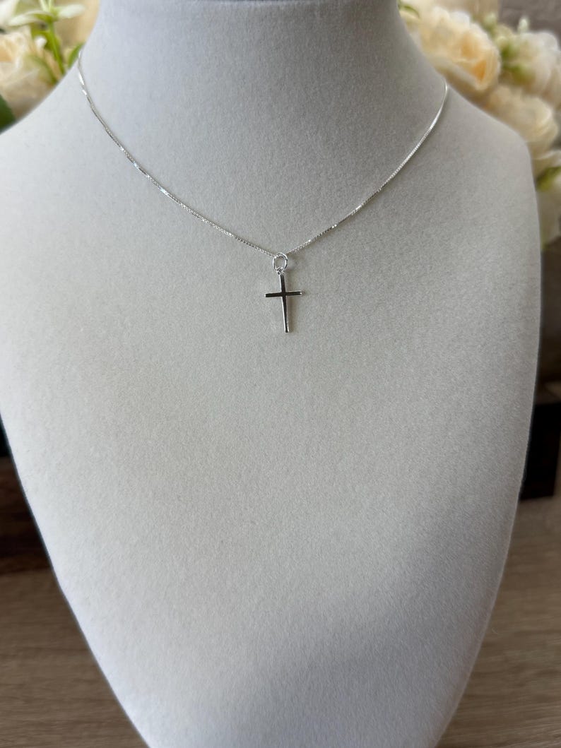 May include: A silver-toned necklace featuring a thin, delicate chain and a small, simple cross pendant. The metal has a polished, reflective finish, and the cross is suspended from a circular jump ring attached to the center of the chain.